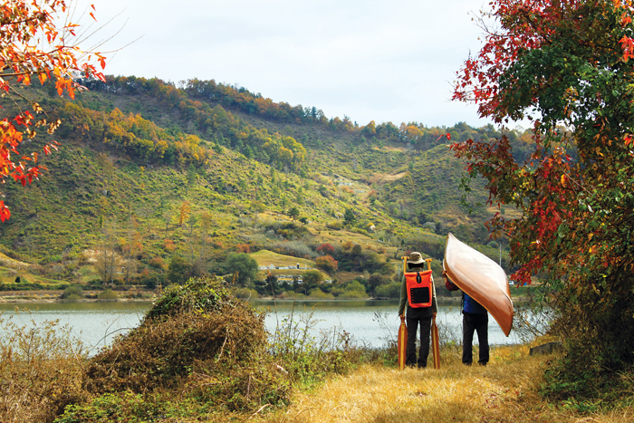 단풍이 아름다운 숲속 강가에 서 있는 두사람, 한사람은 노를 한사람은 카누를 이고 있다