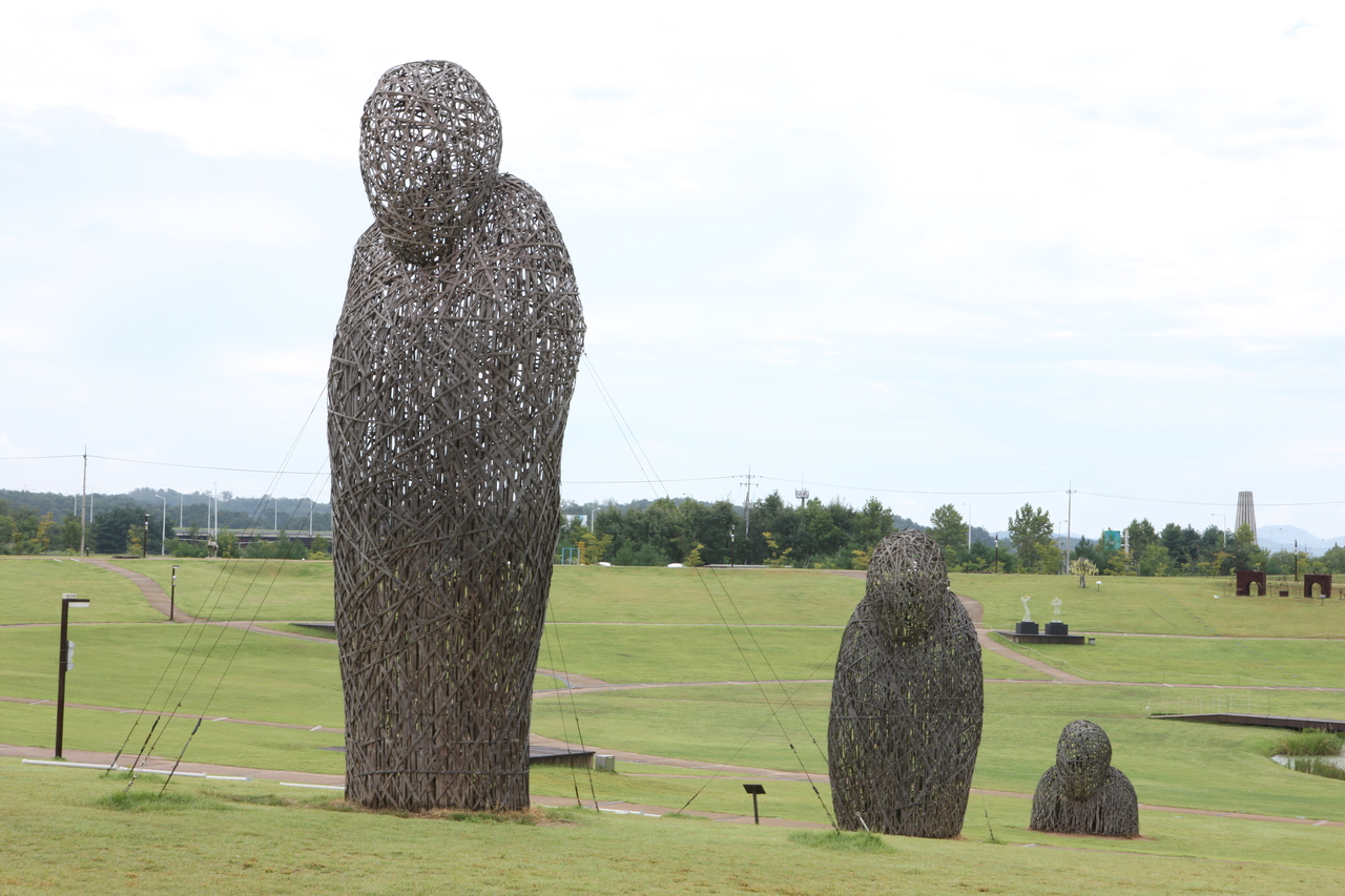 Large woven human-shaped sculptures standing on open green lawns in a public park