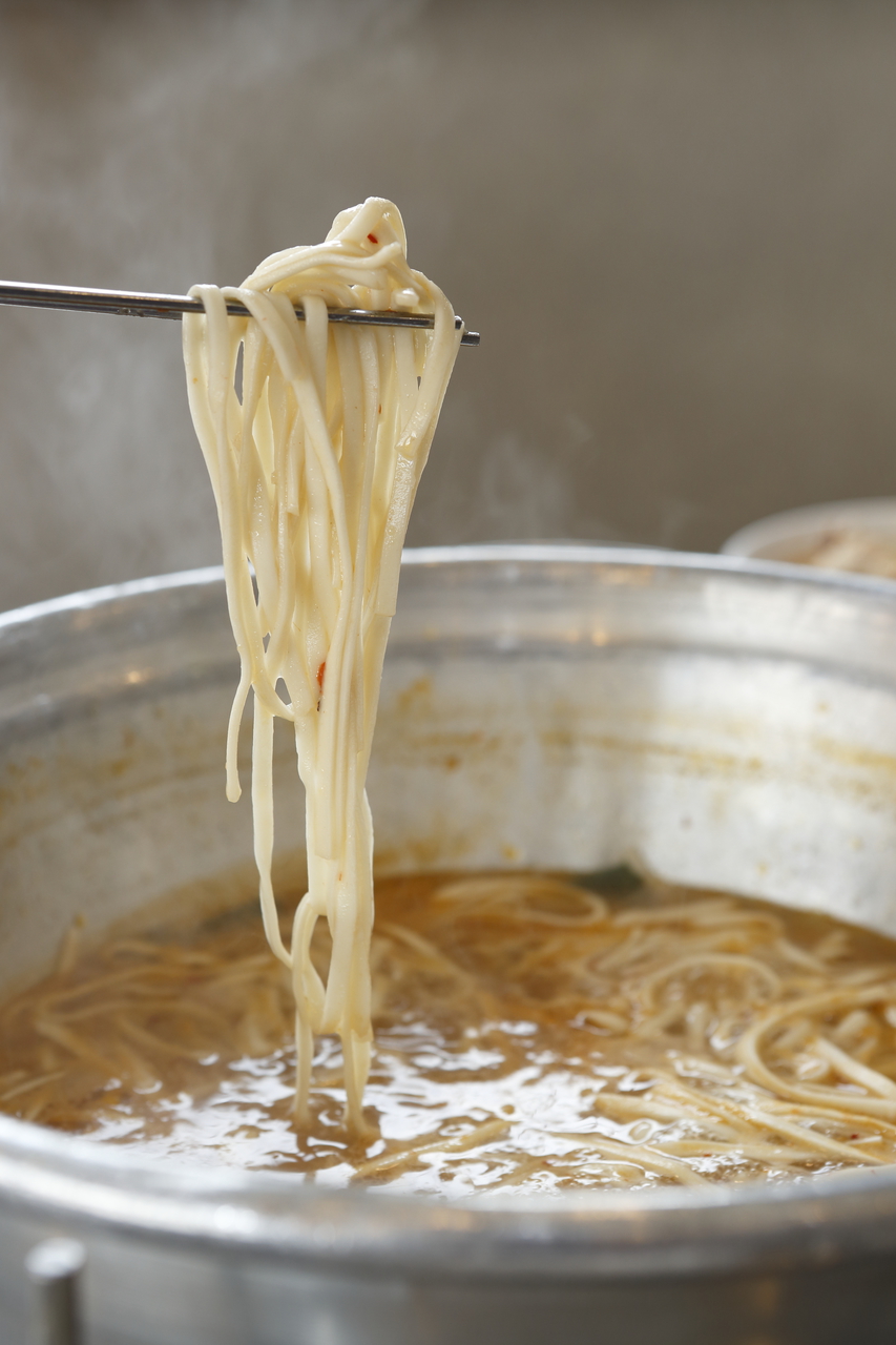 Wheat noodles lifted with chopsticks from a steaming metal pot of chicken broth