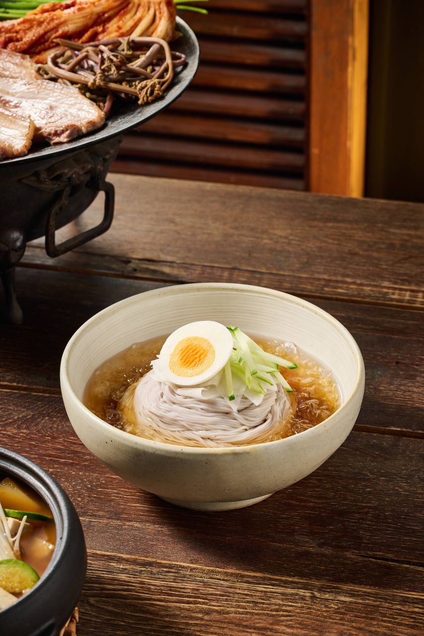 A bowl of mul-naengmyeon, cold buckwheat noodles in icy broth with egg, beside a grill