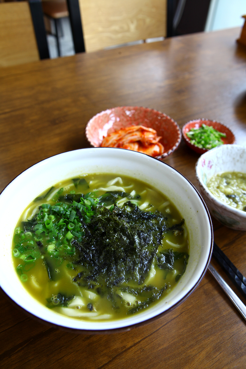 A bowl of kalguksu, knife-cut noodles in broth with seaweed and scallion, with kimchi on the side